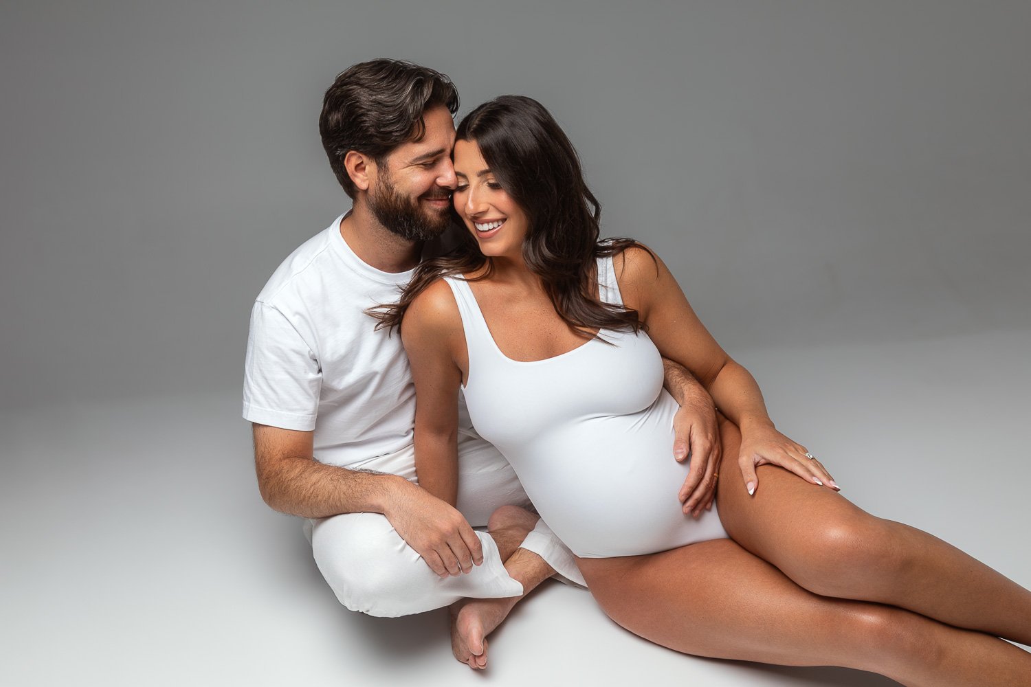 A pregnant woman and a man sit together, smiling and touching her belly, against a plain gray background. maternity photoshoot.

