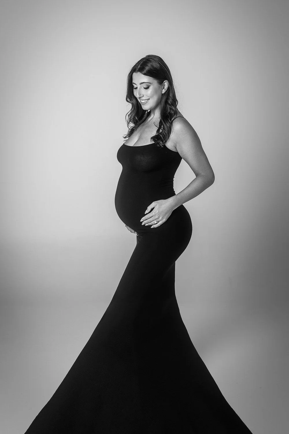 Black and white maternity photoshoot portrait of a pregnant woman in a sleeveless black dress, smiling and looking down, standing against a plain background.
