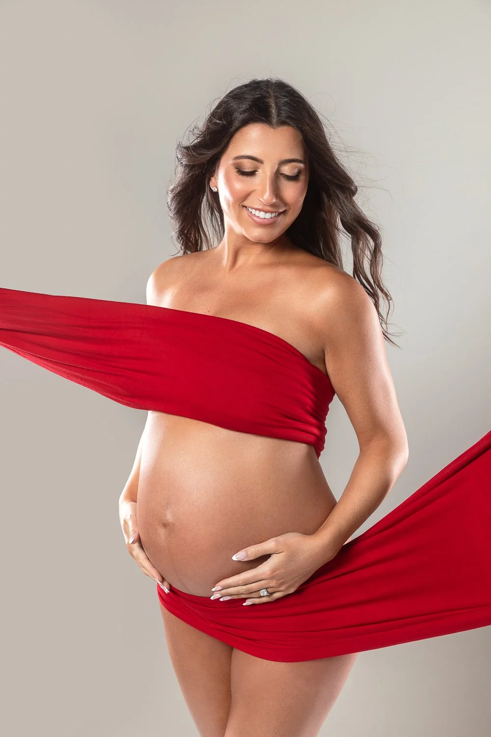 A pregnant woman with long dark hair smiling, wearing a red fabric draped around her chest and waist, standing against a light gray background. maternity photoshoot.