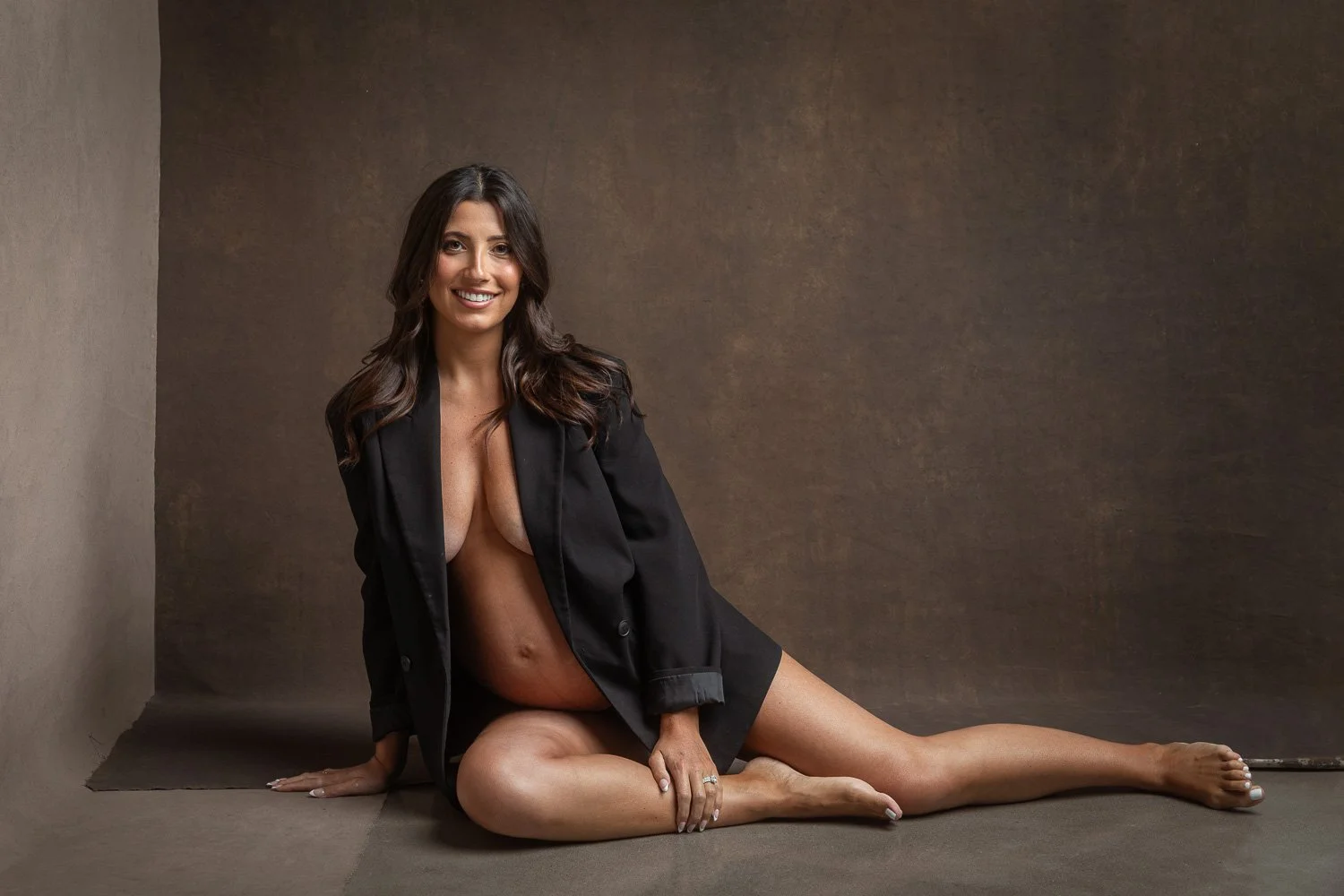 A woman with long dark hair, sitting on the floor with one leg extended and the other bent, wearing a black blazer and no shirt, smiling at the camera against a plain brown background. maternity photoshoot.