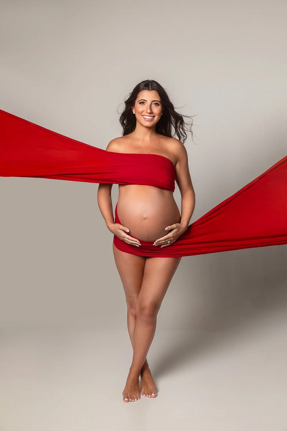 Pregnant woman with long dark hair smiling at camera, wrapped in red fabric covering her chest and hips, standing barefoot against a neutral background. maternity photoshoot.