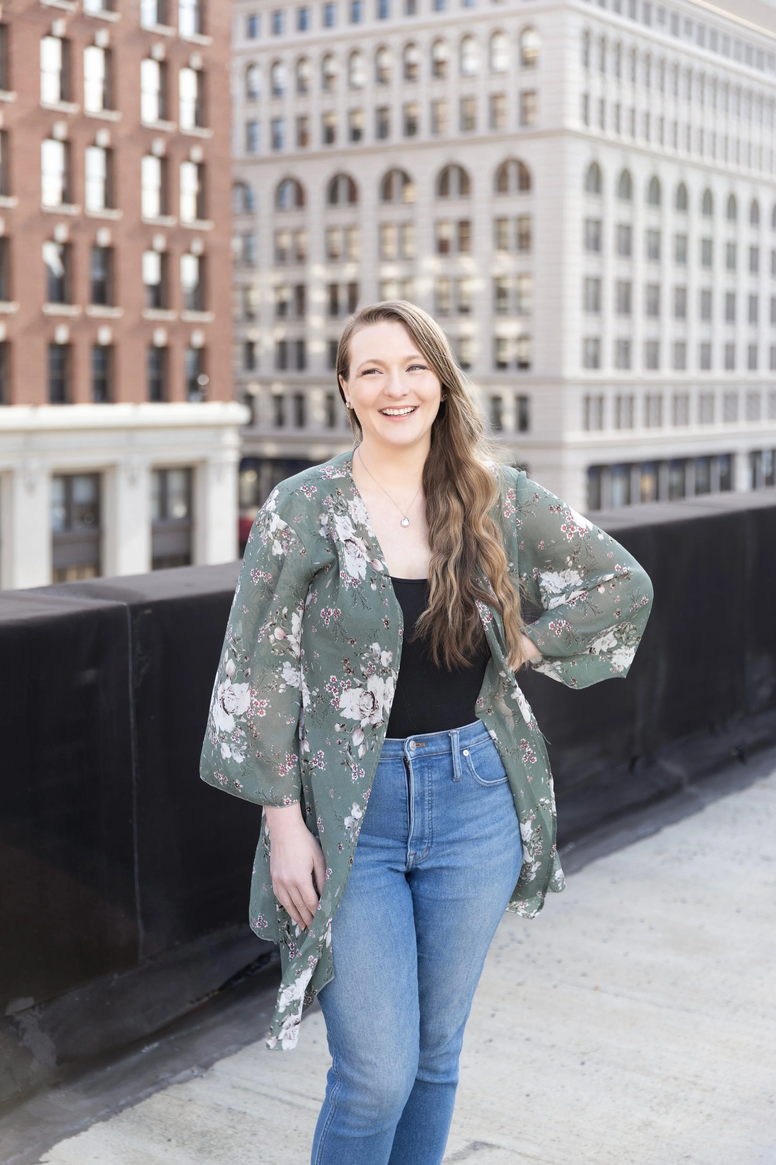 A young woman with long wavy hair smiling outdoors on a rooftop in front of tall buildings.