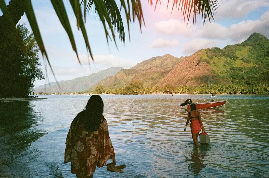 Two women walking in shallow water near a boat with lush green mountains in the background.