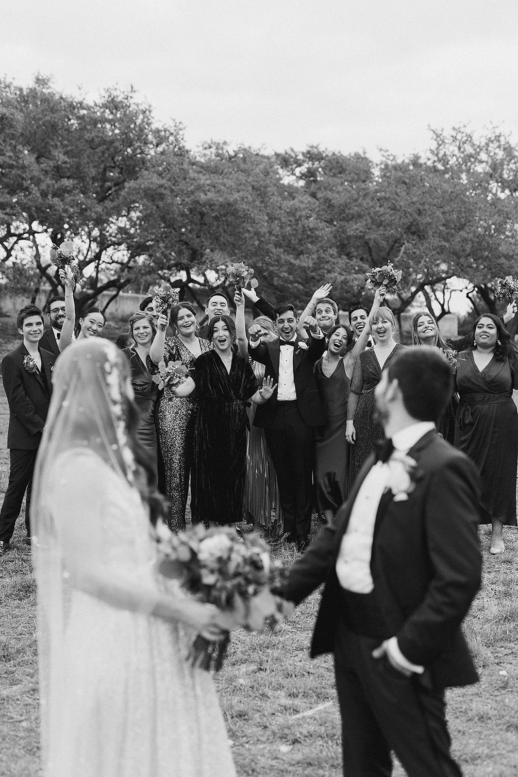 Black and white photo of a wedding scene with a bride and groom in the foreground, gazing at each other, while a group of guests cheer and celebrate in the background outdoors with trees.