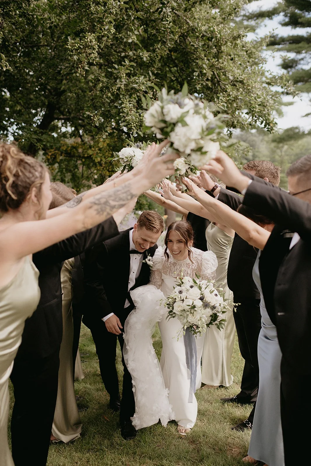 A bride and groom smiling as they walk through an arch formed by wedding guests holding bouquets outdoors.
