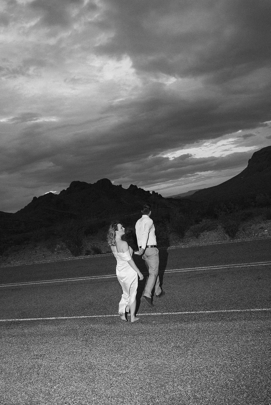 A couple holding hands and walking on an empty road with mountains and cloudy sky in the background, black and white photograph.