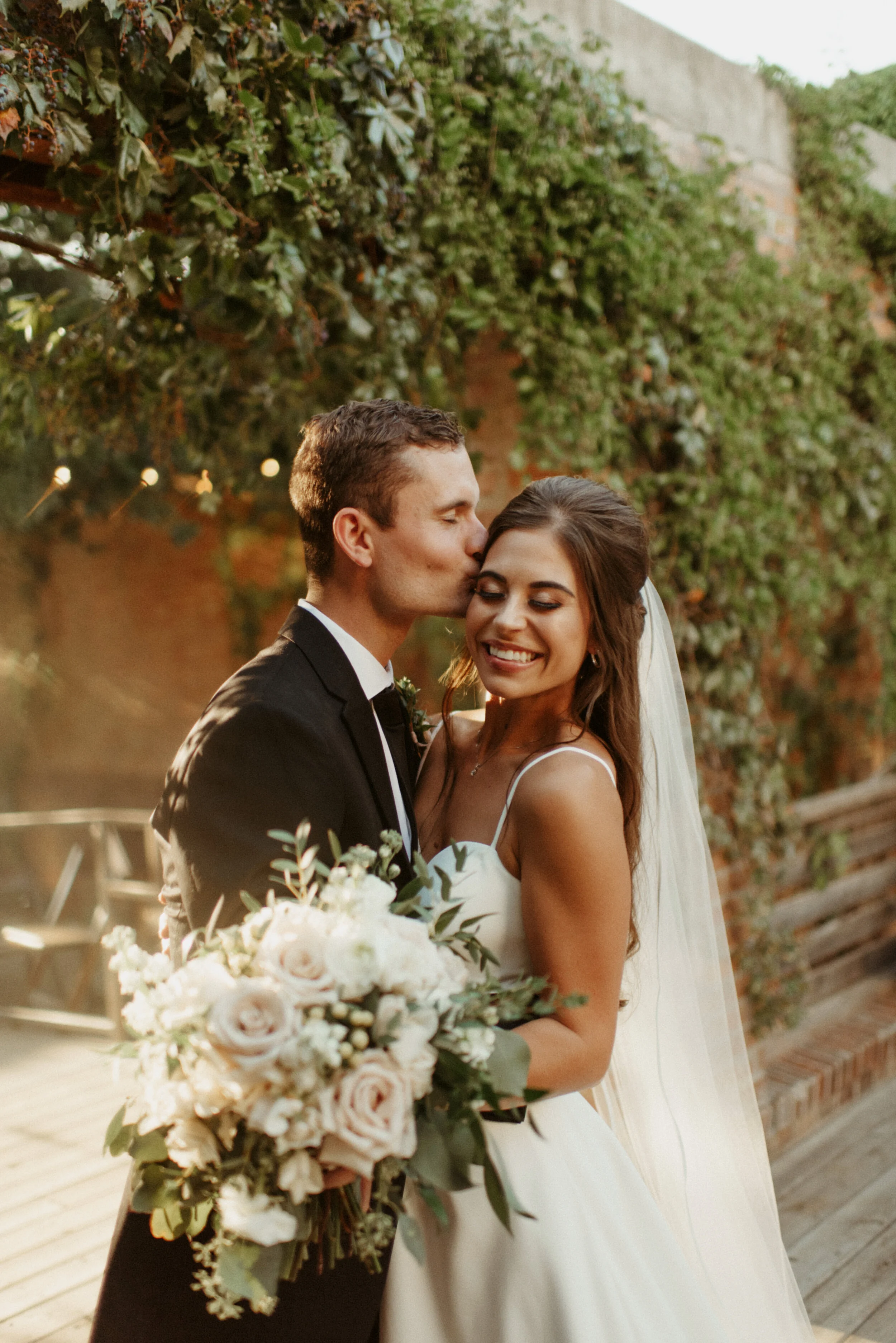 A bride and groom share a kiss outdoors, the bride holds a large bouquet of white and blush roses with greenery, the groom wears a black tuxedo, and the scene is lit with warm, natural sunlight.