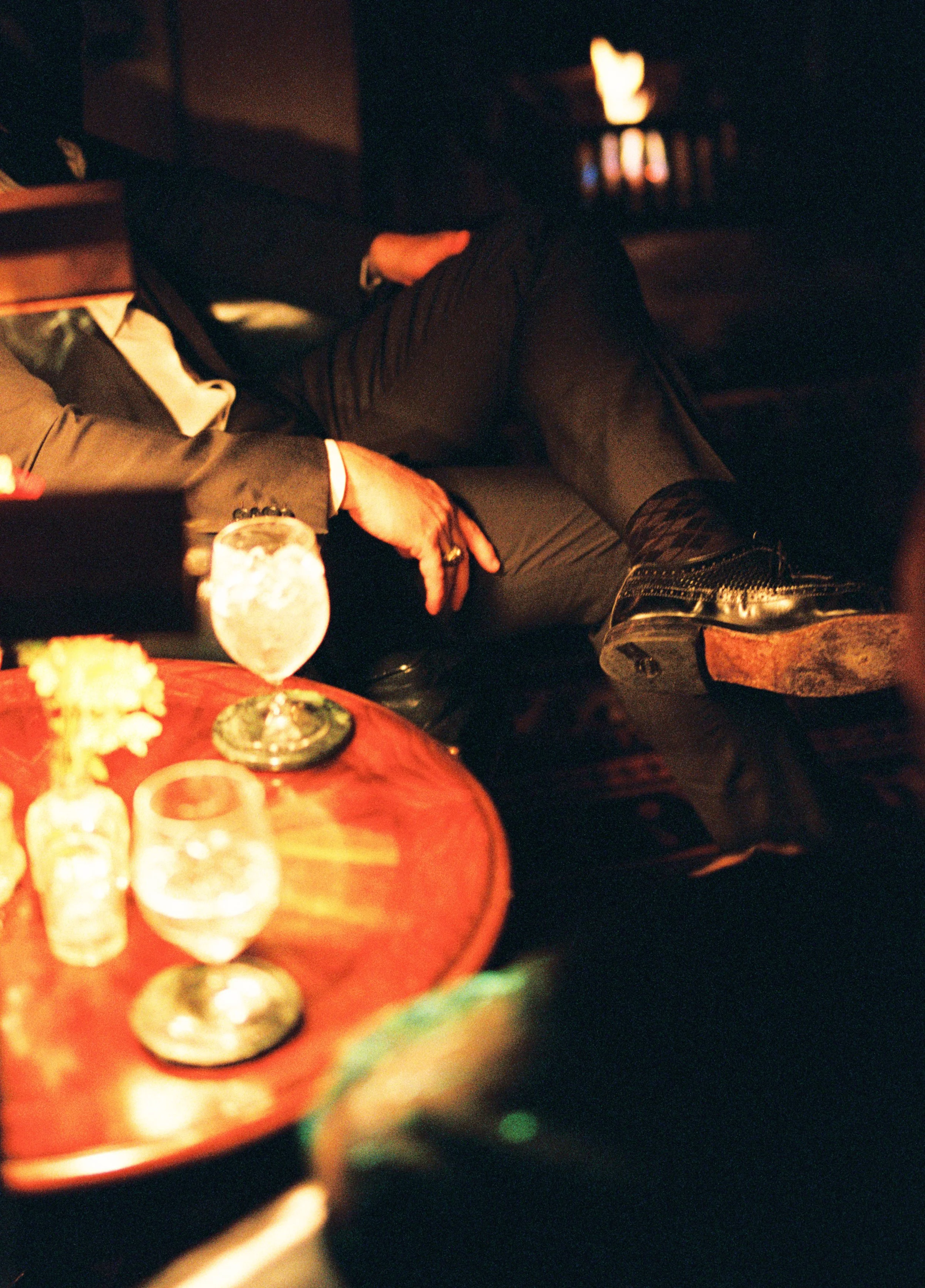 A person dressed in a suit sitting comfortably with legs crossed in a dimly lit lounge or bar setting. On a wooden table in the foreground, there are wine glasses, a small flower vase, and candles or small decorative items.