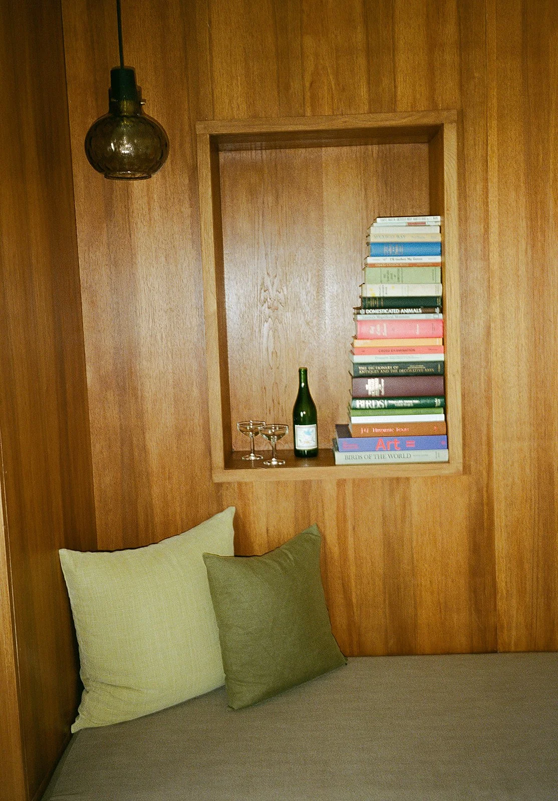 A wooden-walled nook with a built-in shelf holding a bottle of wine, books, and two wine glasses. A bed with two pillows is in the foreground. An overhead light fixture is visible.