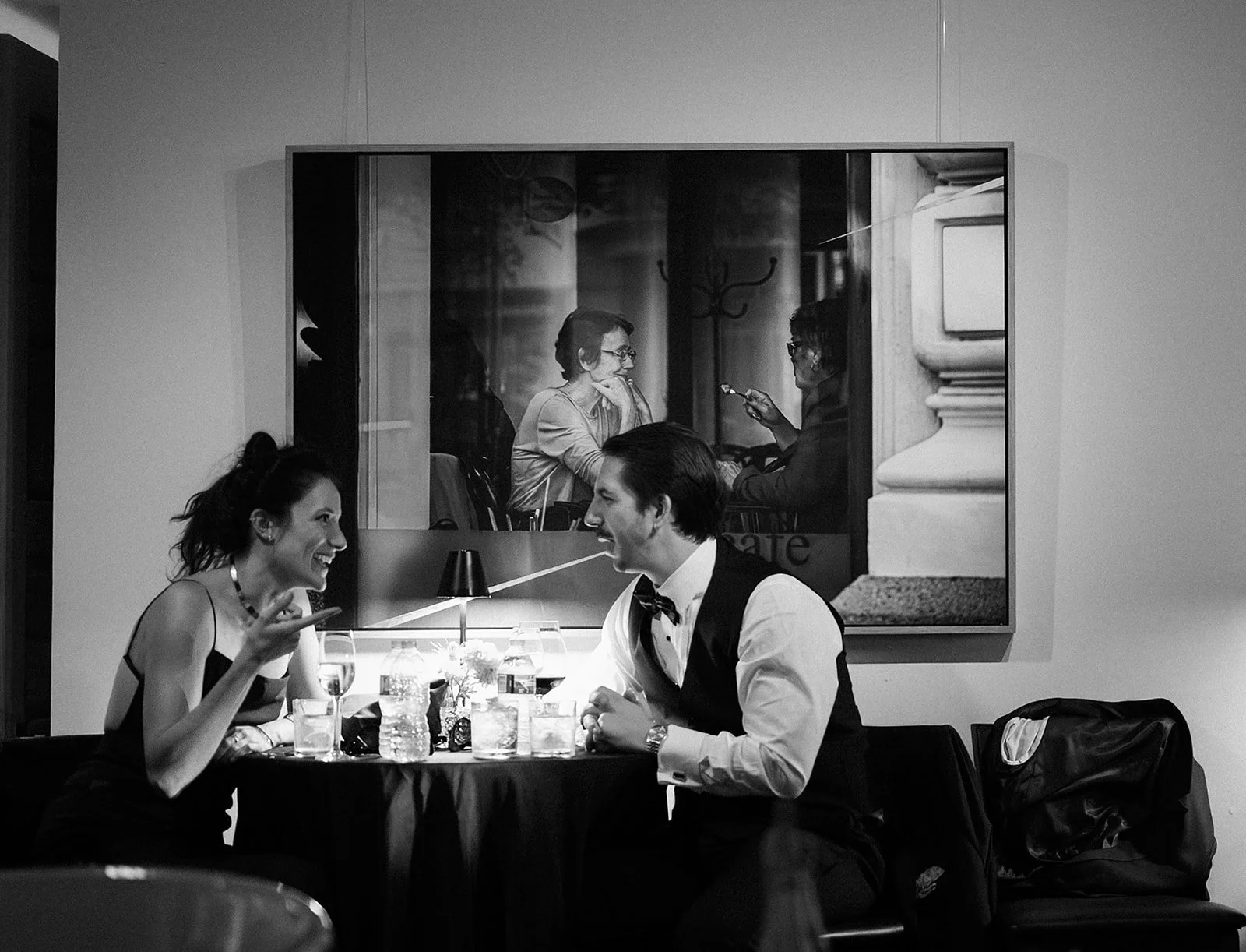 A woman and a man in formal attire sitting at a restaurant table, smiling and engaged in conversation, with a large window behind them showing a reflection of two women talking.