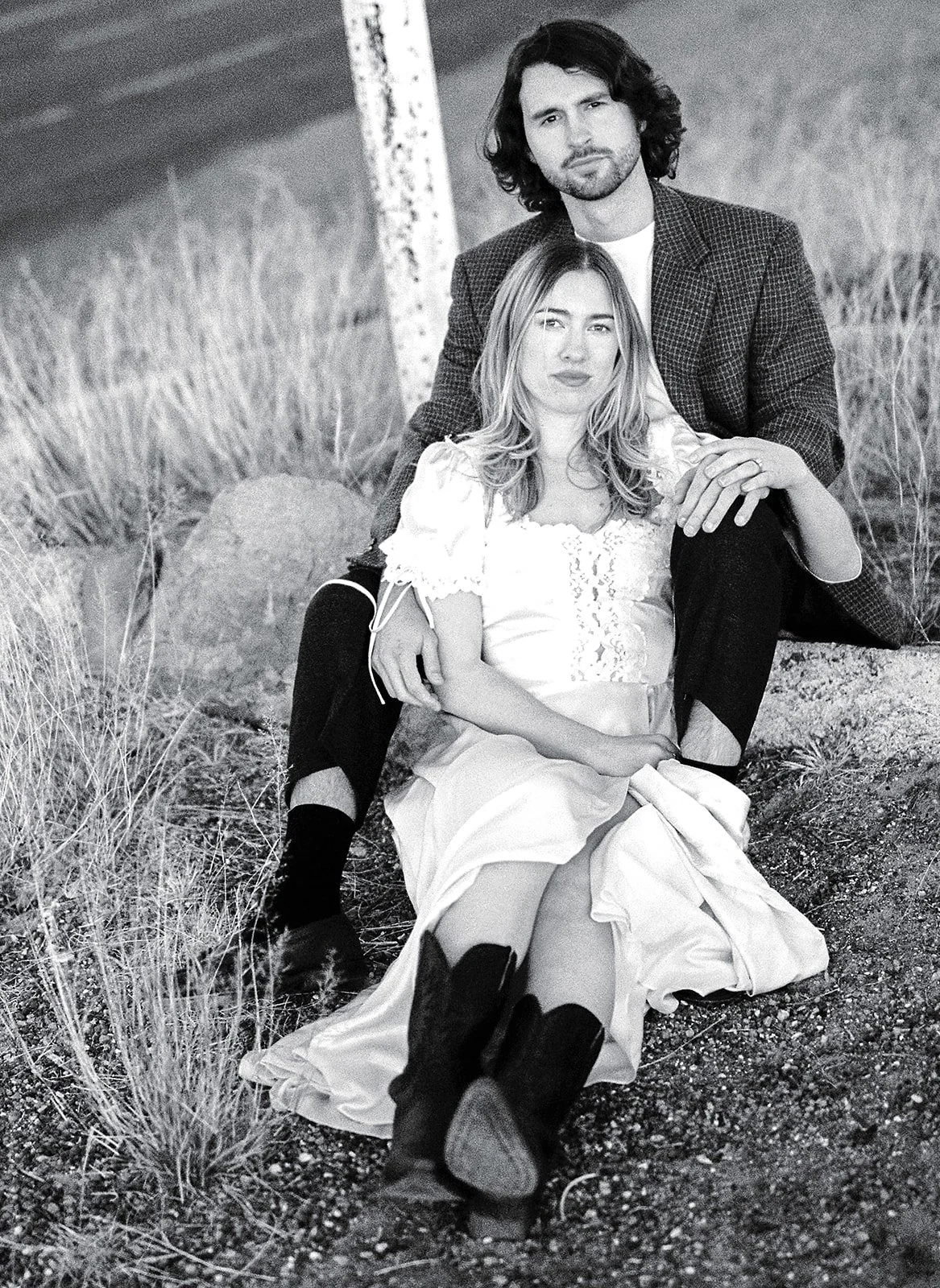 A black and white photo of a man and woman sitting outdoors on rocky ground with tall grass. The man has long curly hair and a beard, wearing a blazer, while the woman has long hair, wearing a white dress and black knee-high boots.