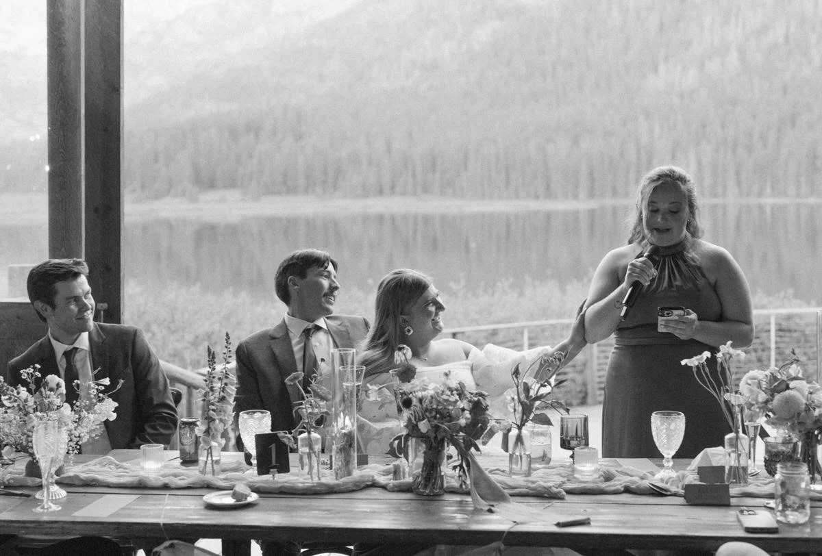 Wedding reception, people at a long table, woman giving speech, floral arrangements, lake view in background.