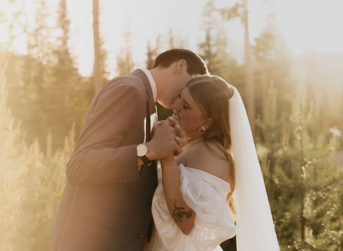 A couple sharing a tender moment during their wedding photo shoot in a forest setting with golden sunlight. The bride wears a white off-shoulder dress and the groom a suit. They are holding hands intimately.