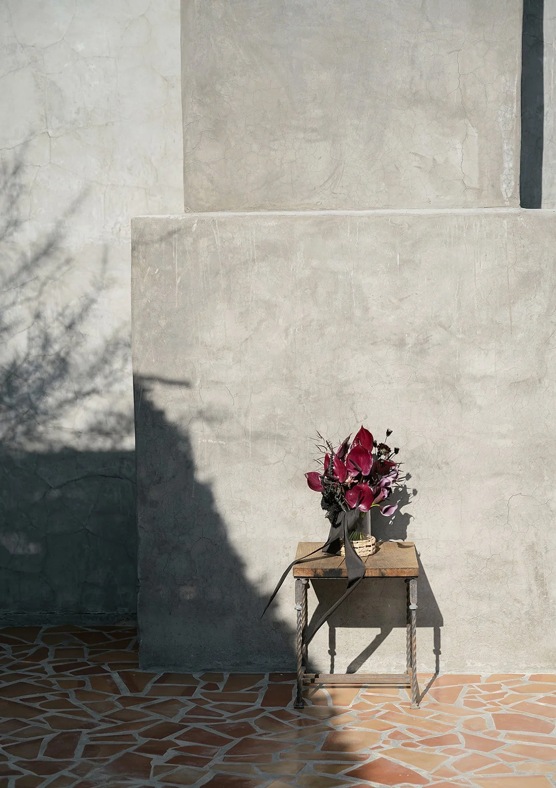A rustic wooden stool with a bouquet of dark red flowers against a textured concrete wall, casting long shadows on a tiled floor.