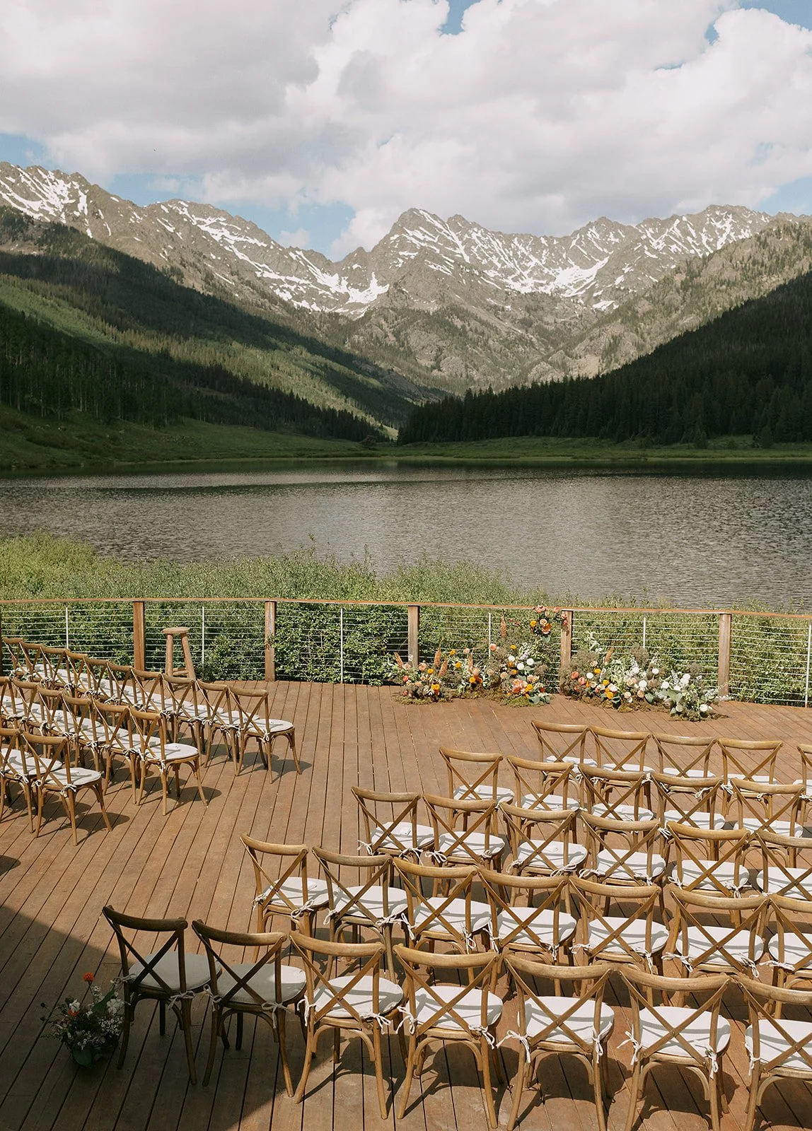 Outdoor wedding ceremony setup with wooden chairs on a wooden deck overlooking a lake and mountainous landscape.
