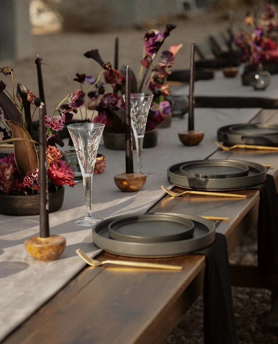 Elegant outdoor table setting with dark plates, gold cutlery, black candles, and floral centerpieces featuring purple and red flowers.
