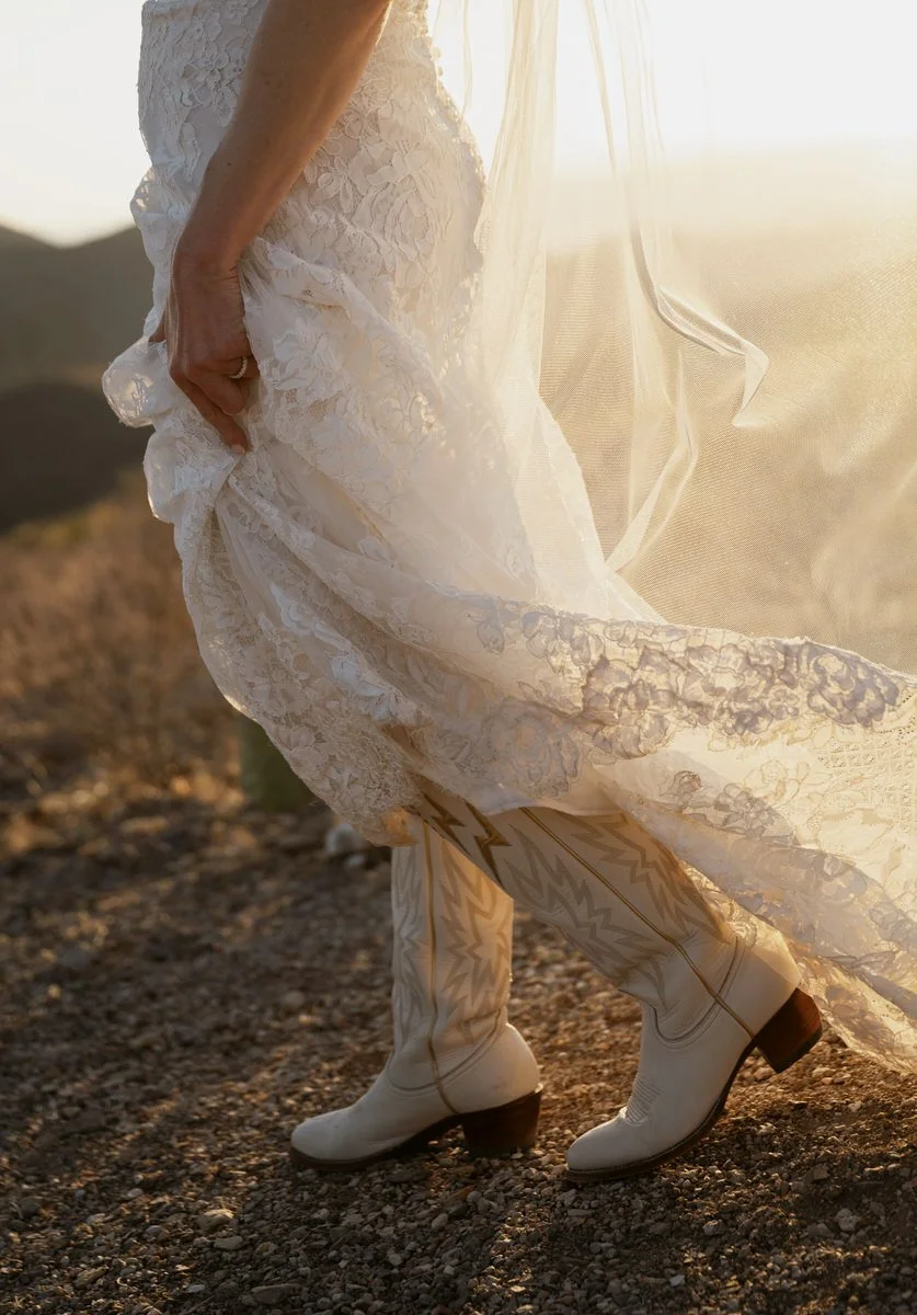 Bride wearing lace wedding dress and cowboy boots standing outdoors with sunlight.
