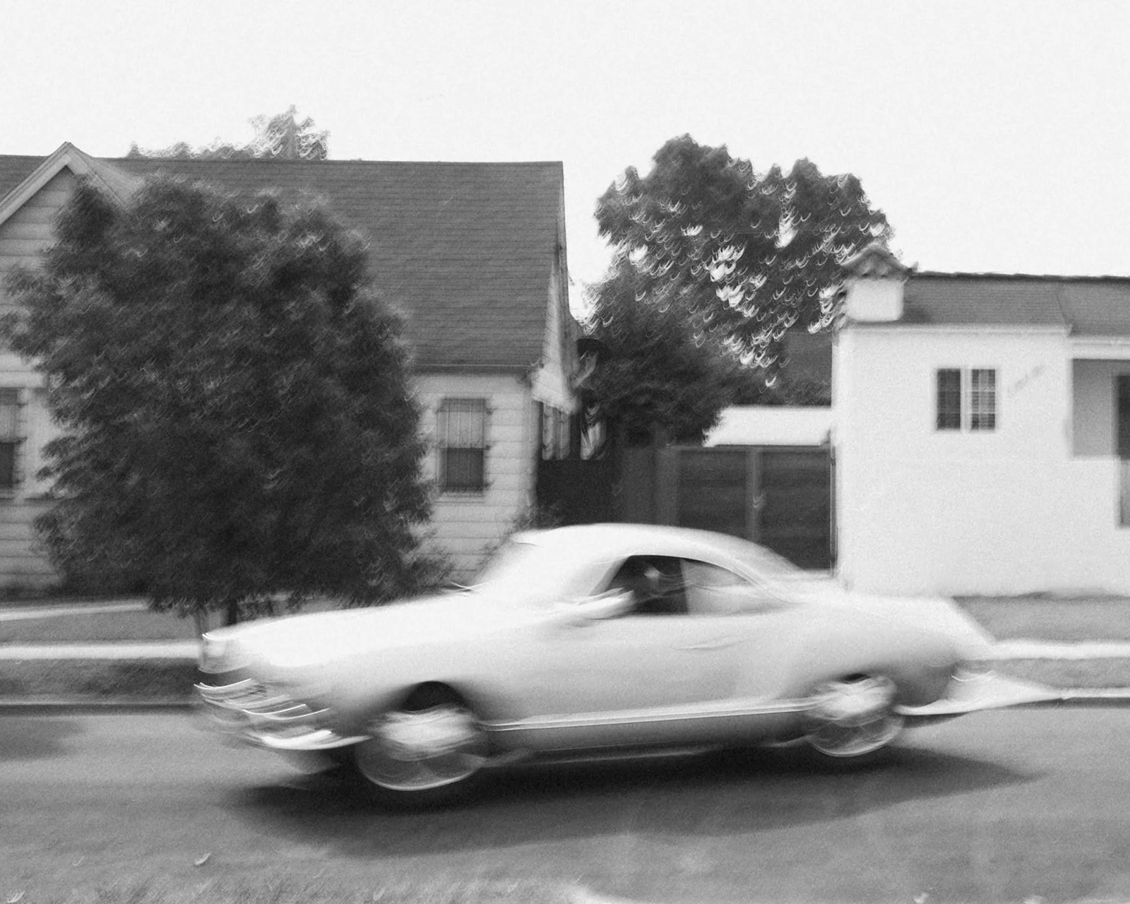 Blurry black and white image of a vintage car driving past suburban houses.
