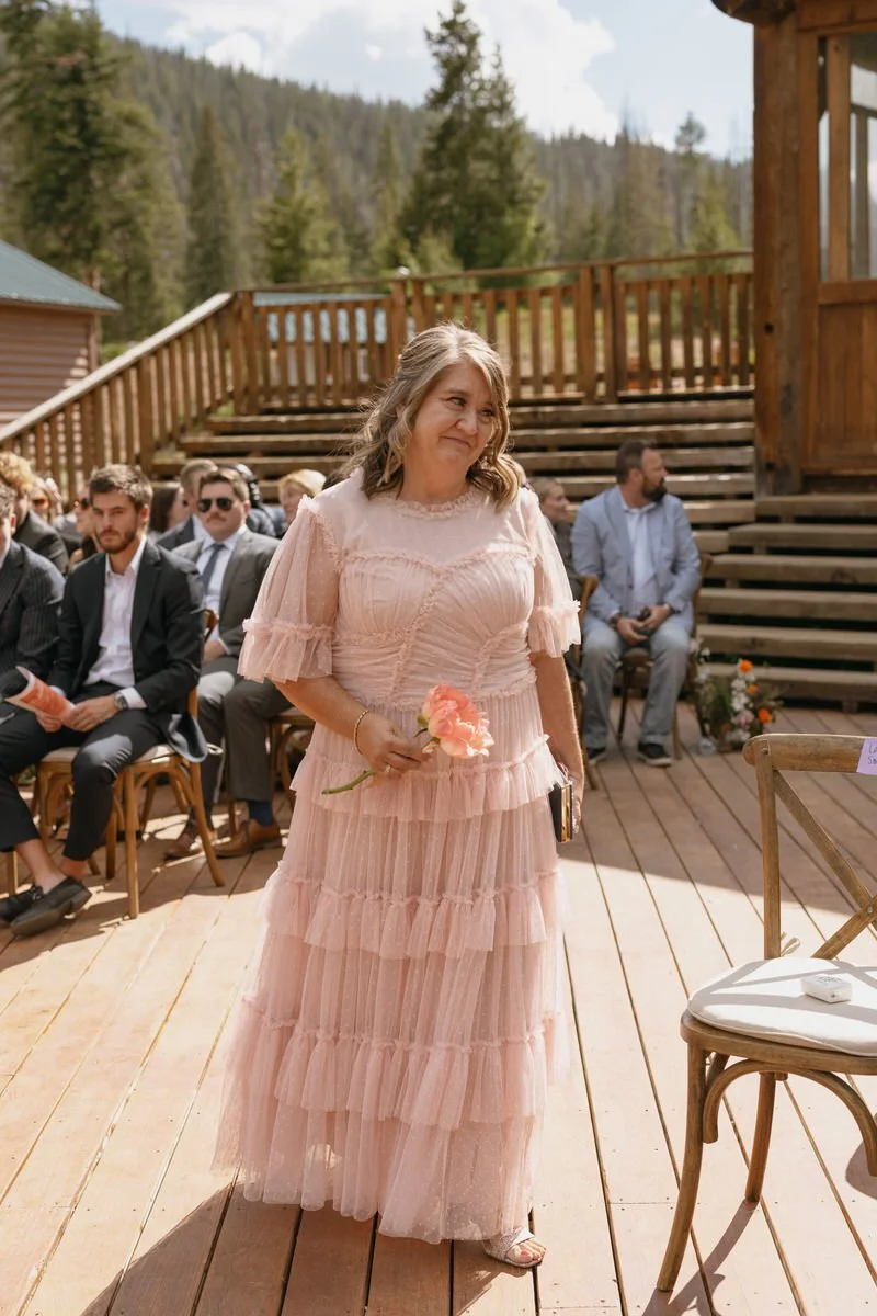 Outdoor wedding ceremony with a woman in a pink ruffled dress holding a flower, walking down a wooden aisle. Seated guests in formal attire are visible, with a forest backdrop.