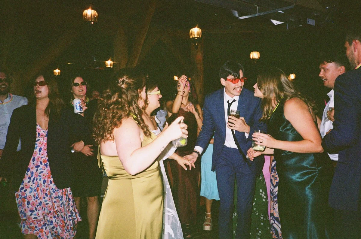 People dancing and socializing at a party, dressed in formal attire and wearing sunglasses, holding drinks.