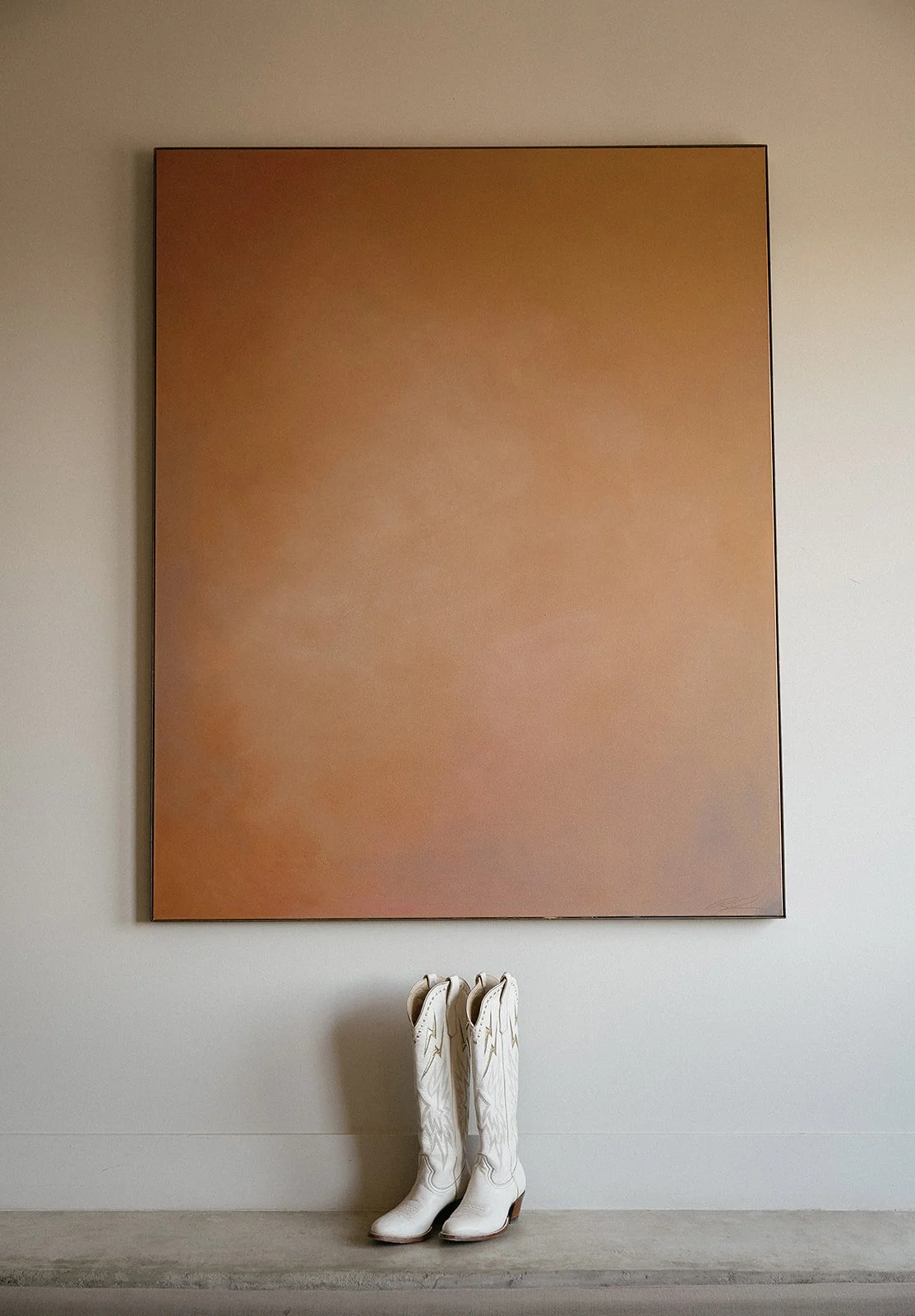 Pair of white cowboy boots in front of a large abstract brown painting.