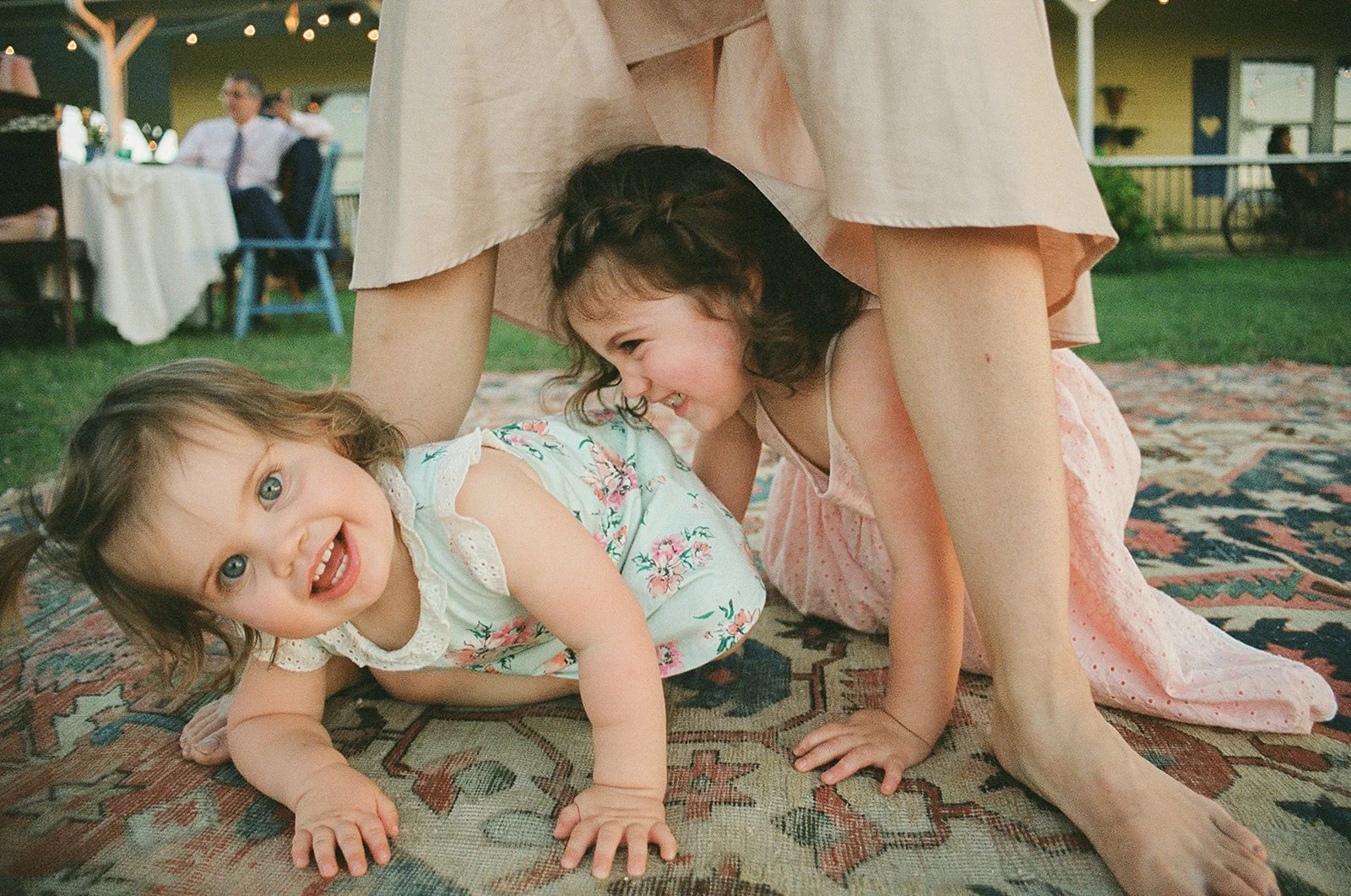 Two young children playing on a patterned rug outdoors. One child is lying down and smiling, while the other is crouched beside them, also smiling. They are under an adult's dress, which partially covers them.
