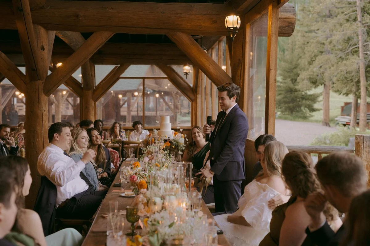 A wedding reception held in a rustic wooden lodge. Guests are seated at a long table adorned with flowers, enjoying a speech from a man in a suit holding a microphone. The atmosphere is warm and celebratory, with a mix of natural and decorative light