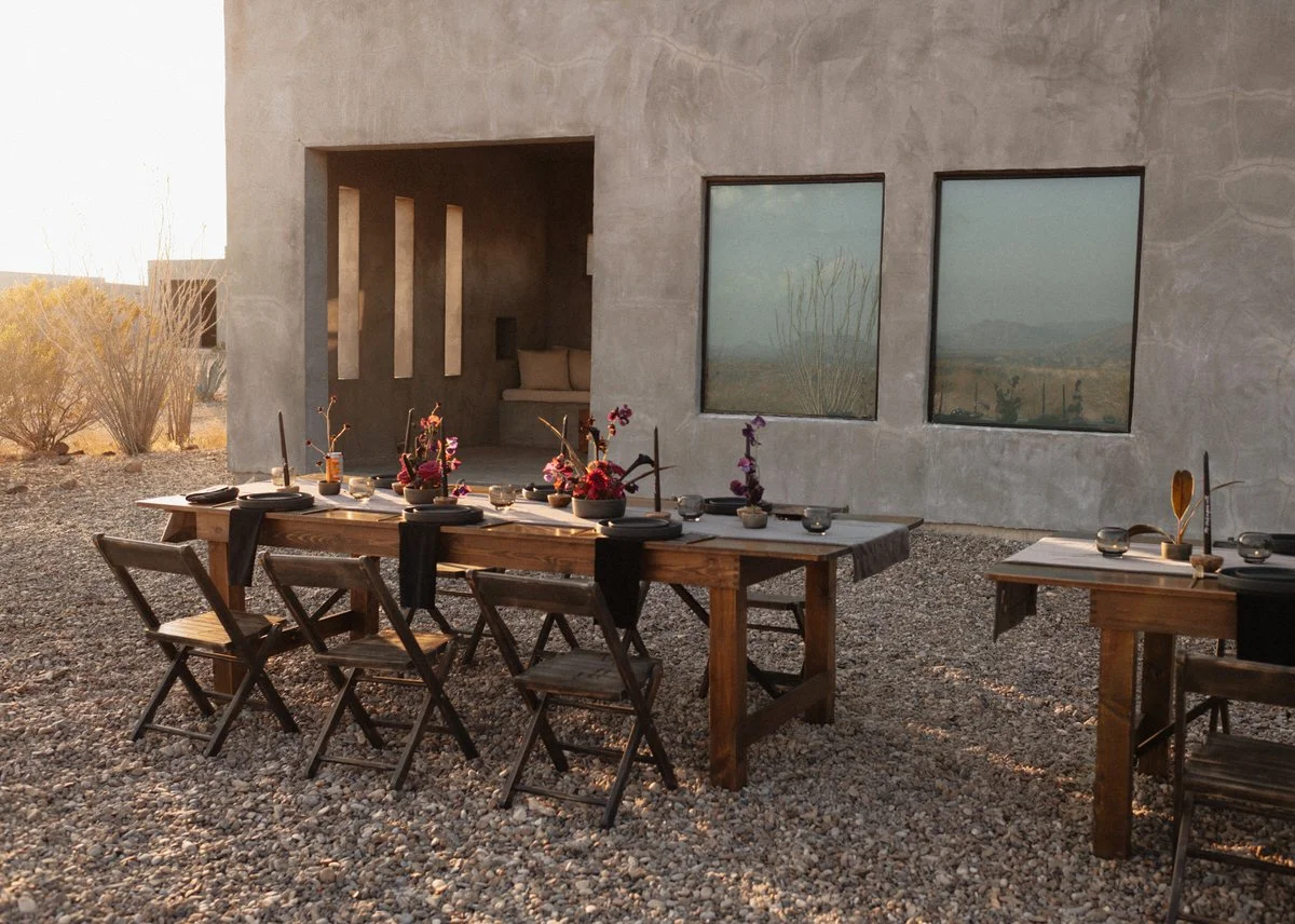 Outdoor dining setup with rustic wooden tables and chairs on gravel surface, adorned with floral centerpieces and place settings, next to a building with large windows.
