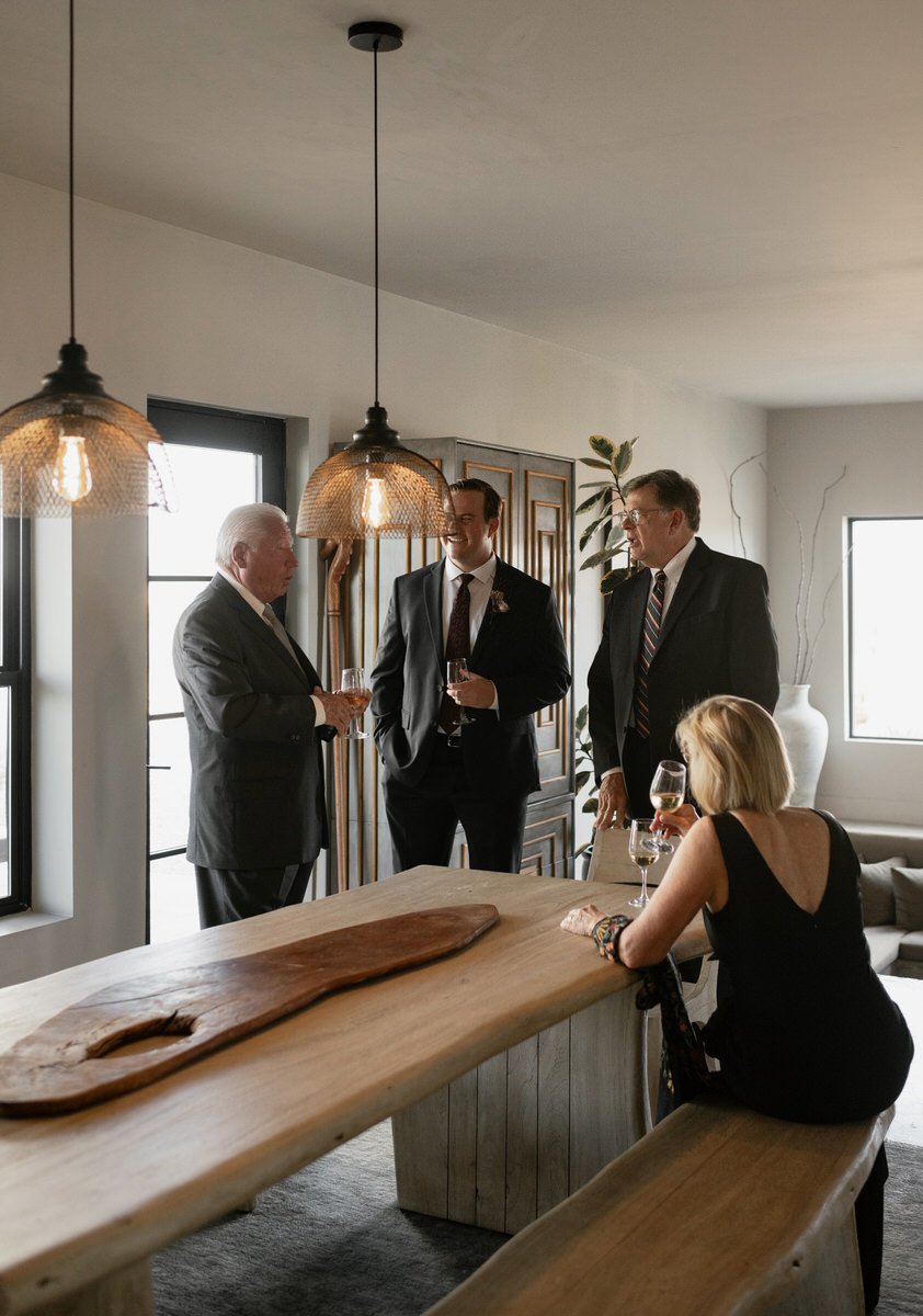 Four adults in formal attire standing and sitting around a wooden table, holding glasses of wine, in a well-lit room with pendant lights and large windows.