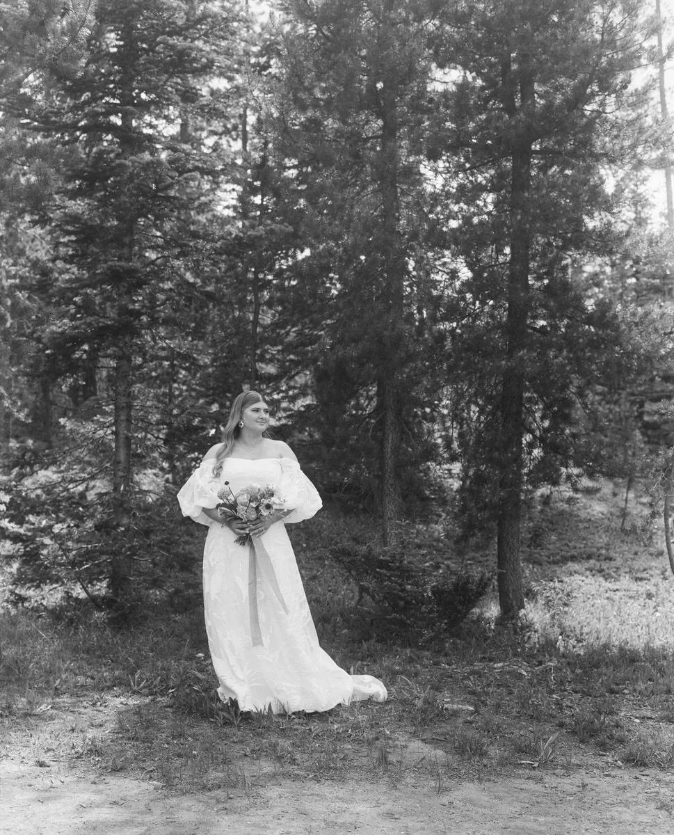 Bride in a wedding dress holding a bouquet in a forest setting, black and white photo.