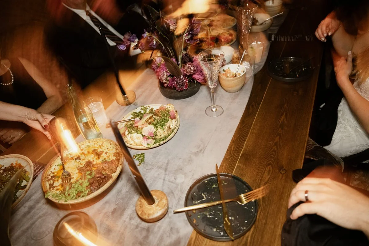 A dimly lit dinner table with plates of food, a floral centerpiece, and candles. There are people seated, partially visible, with one hand reaching for a drink. The focus appears to be on the relaxed dining atmosphere.