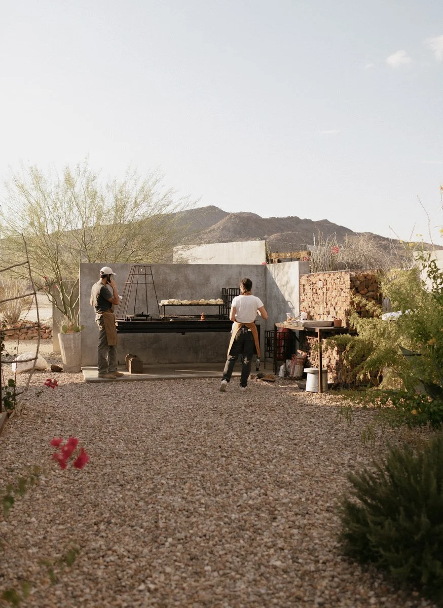 Two people cooking outdoors in a desert setting, standing near a grill with firewood stacked nearby and mountains in the background.