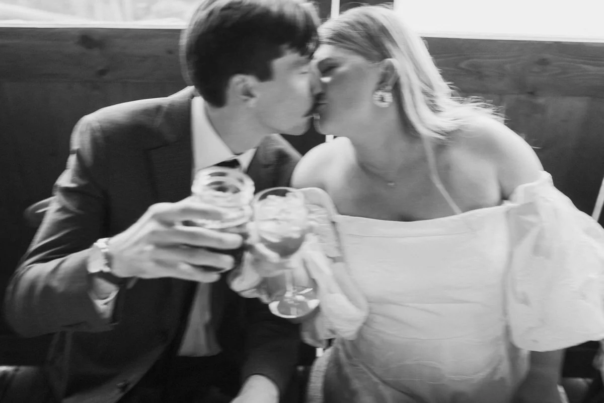 Black and white photo of a couple kissing while holding drinks, with the woman in an off-shoulder dress.