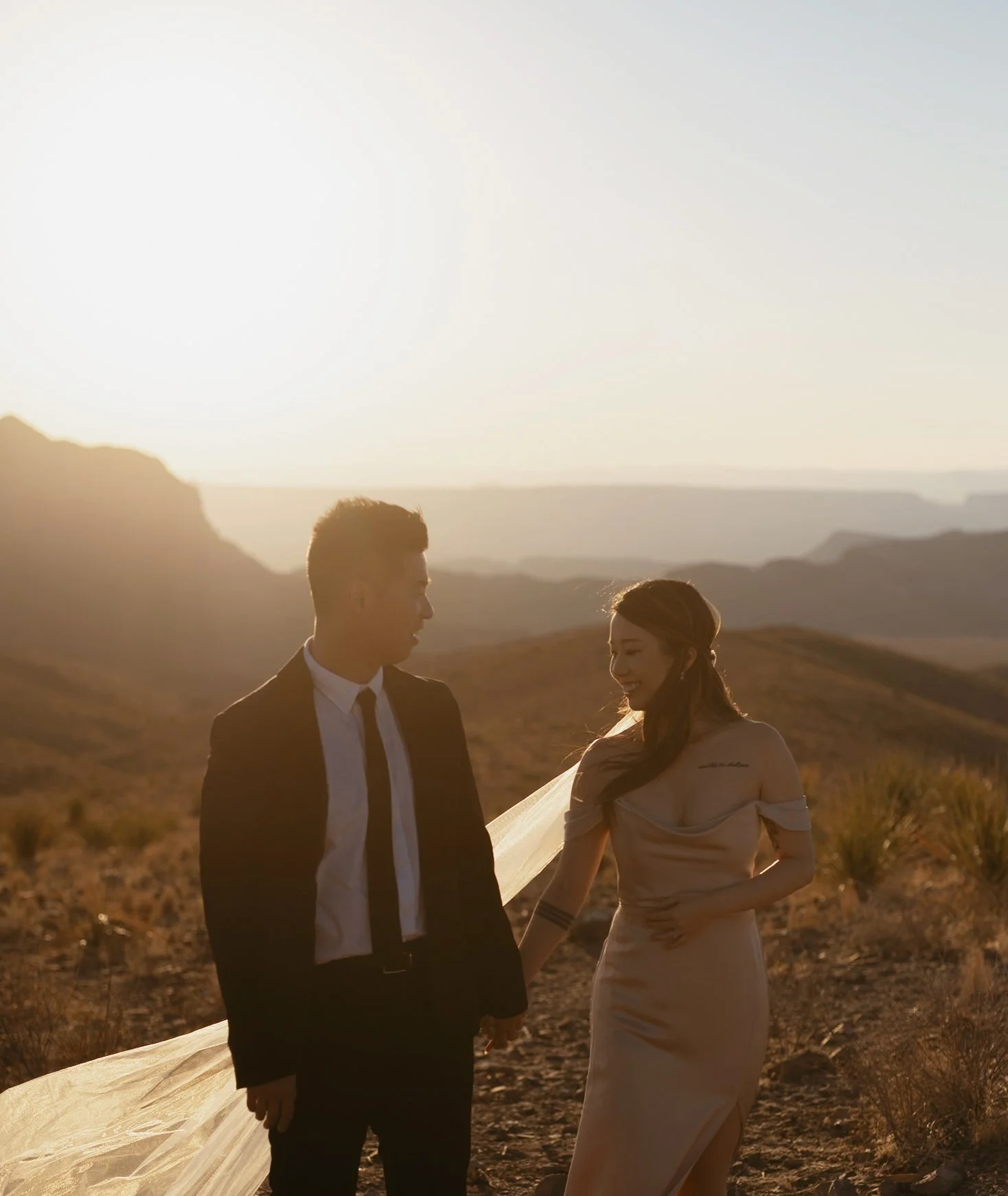 Couple in formal attire walking in a desert landscape at sunset.