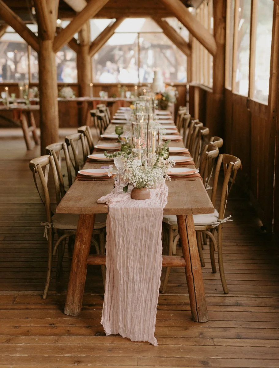 Rustic wooden dining table set for a banquet, adorned with a flowy pink table runner, white floral centerpieces, and elegant table settings, set inside a wooden structure with chairs on each side.
