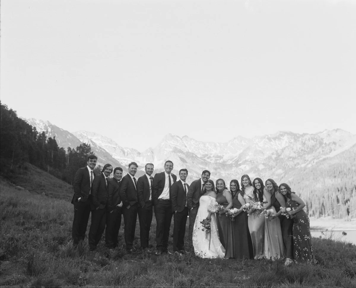 Black and white group photo of people dressed in wedding attire standing outdoors with mountains in the background.