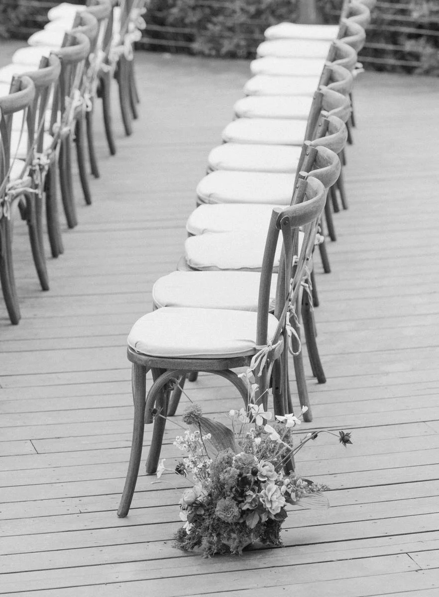 Black and white image of rows of wooden chairs with cushions on a wooden deck, with a bouquet placed on the ground near the front chair.