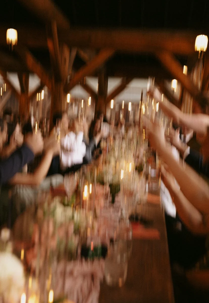 Blurred image of people raising glasses at a banquet table with soft lighting and floral decorations.