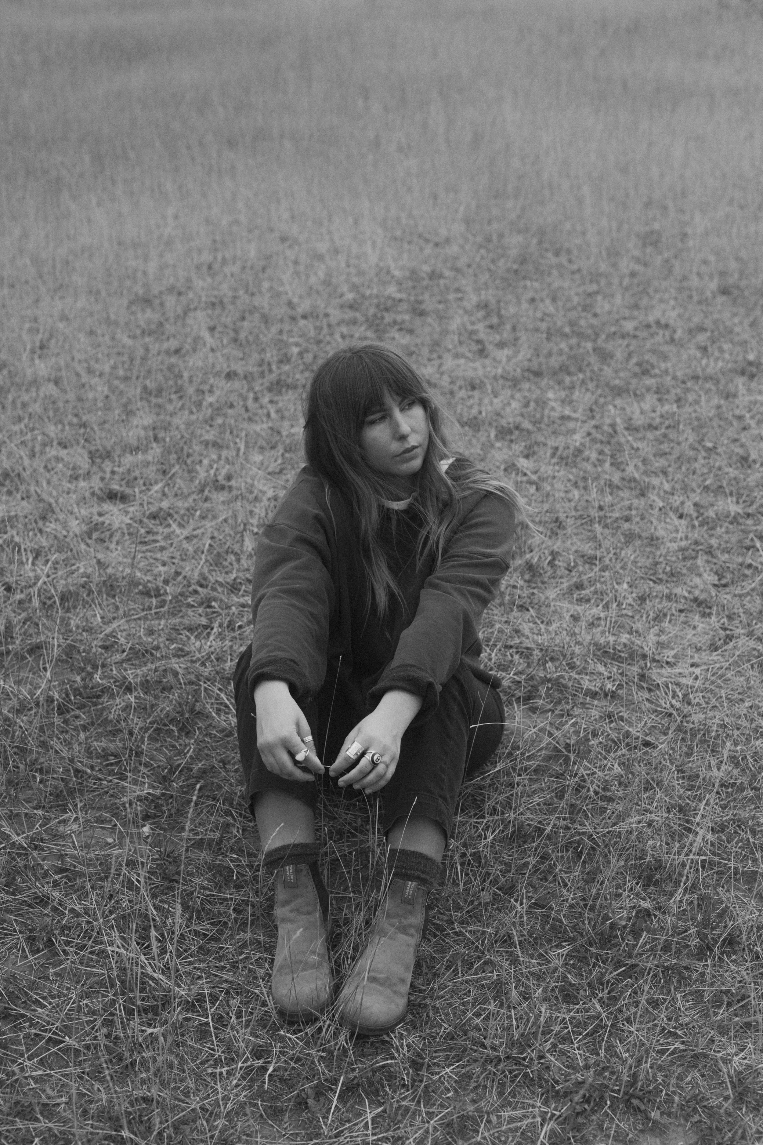 A young woman sitting on the ground in a grassy field, looking contemplative, wearing dark clothing and boots, in a black and white photograph.