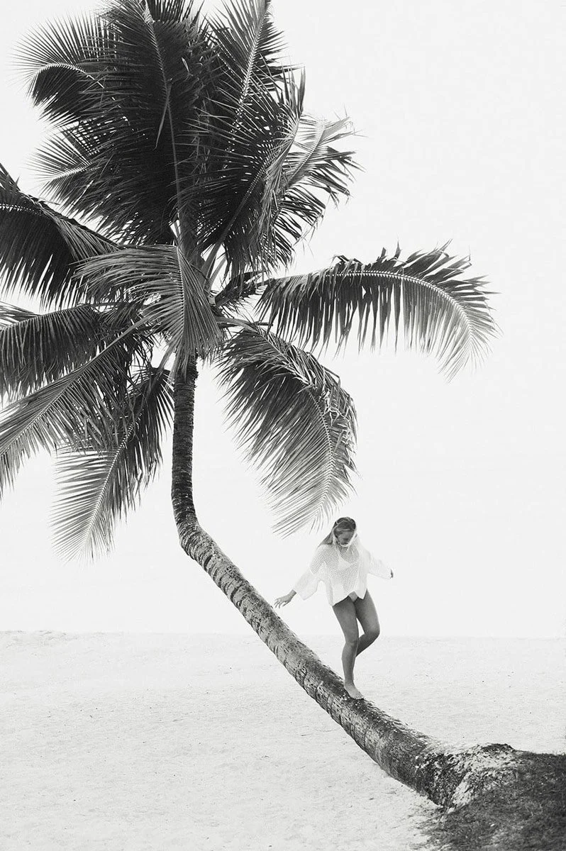 A woman balancing on a leaning palm tree trunk on a beach, with large palm fronds overhead, in black and white.