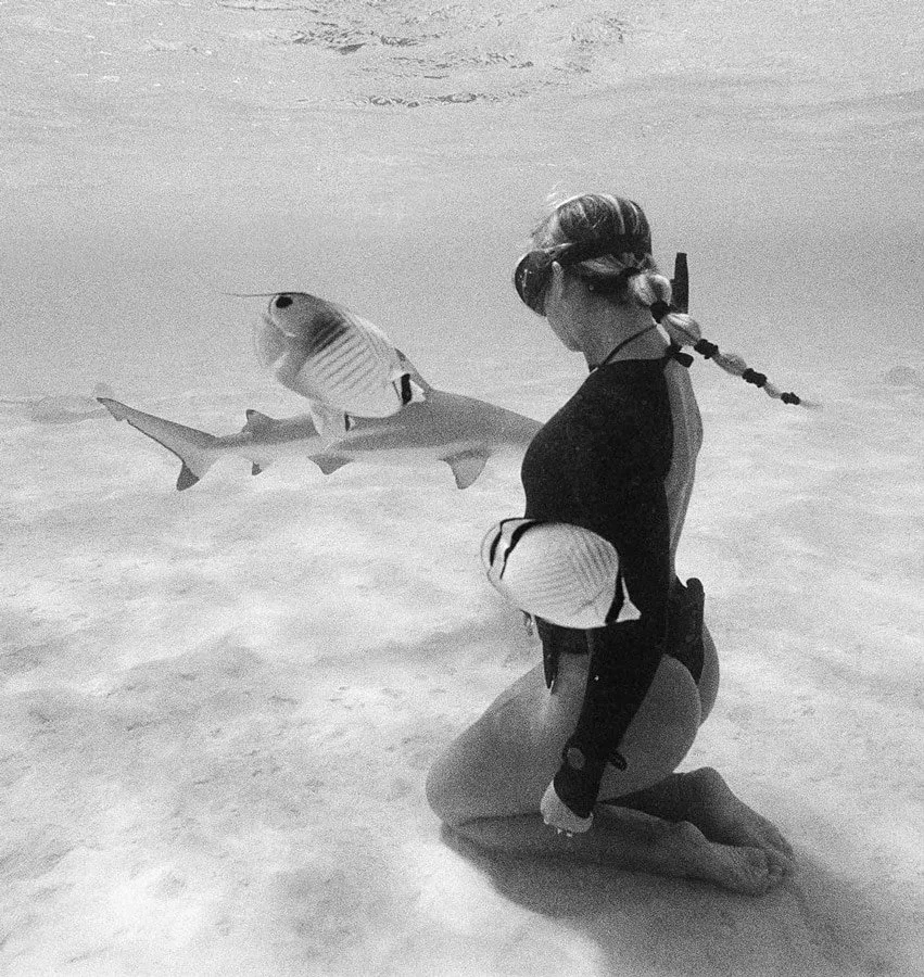 A woman kneels underwater holding a fish in her arms, with a fish swimming nearby.