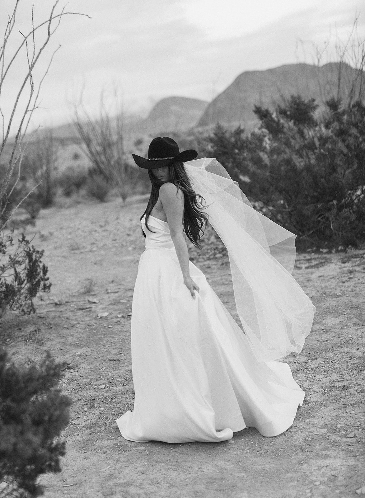 A woman in a black cowboy hat, wedding veil, and wedding dress, walks through the Terlingua desert.