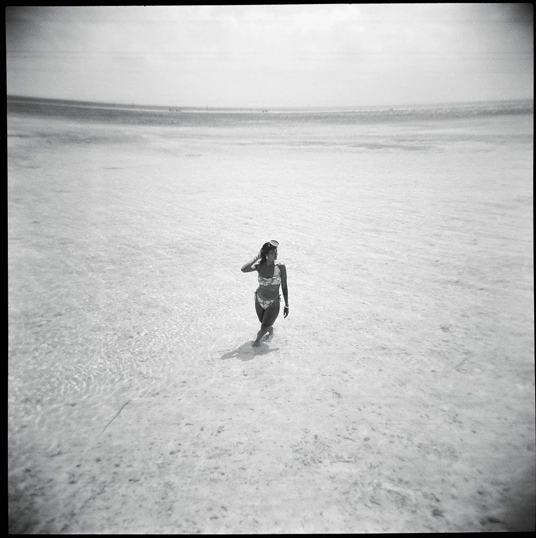 A woman in a swimsuit walking through shallow water at the beach during daytime.
