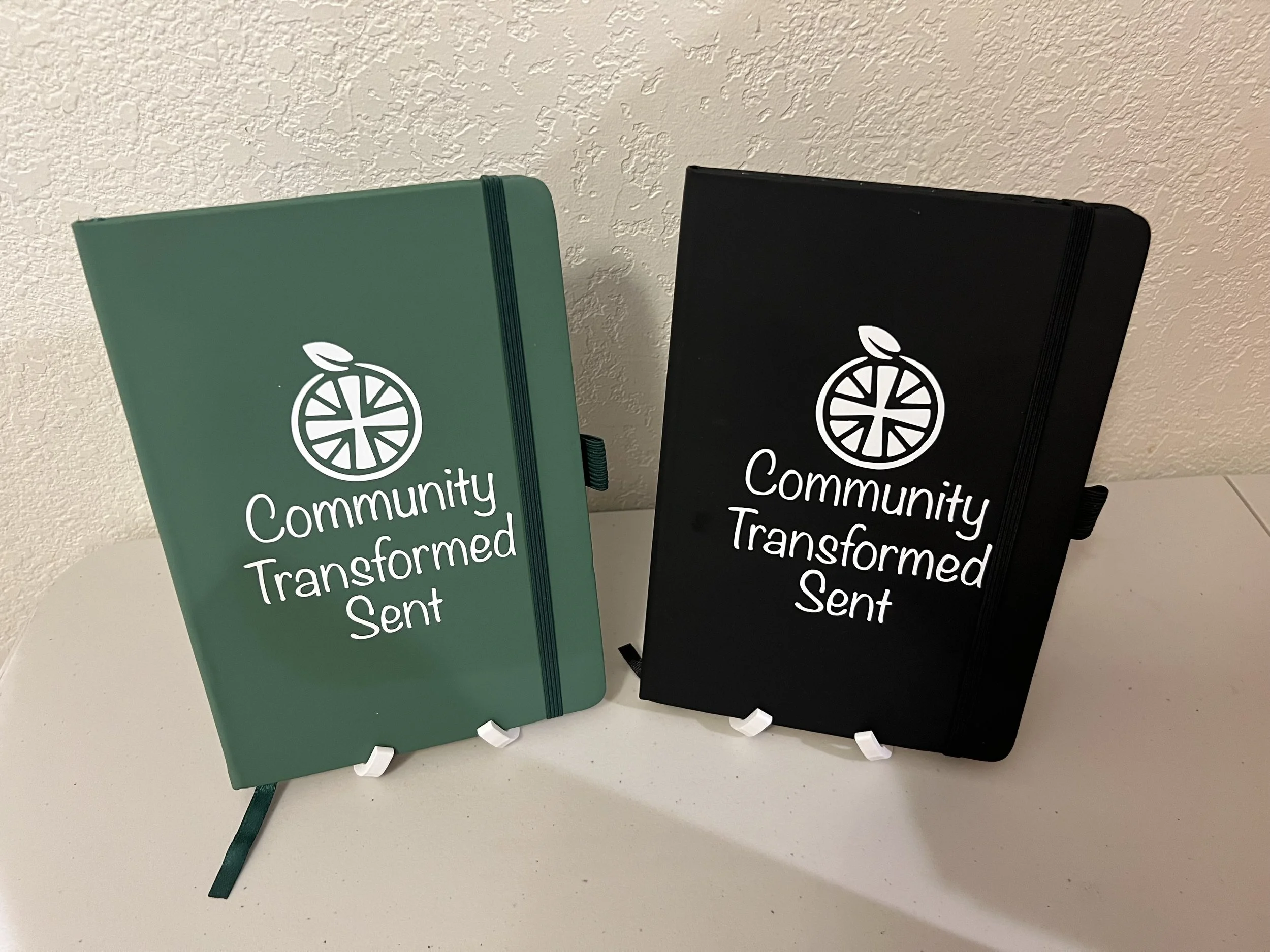 Two notebooks with elastic bands, one green and one black, standing upright against a textured wall. The notebooks have a white logo of a stylized citrus fruit and the word "Community" and "Transformed Sent" printed below.