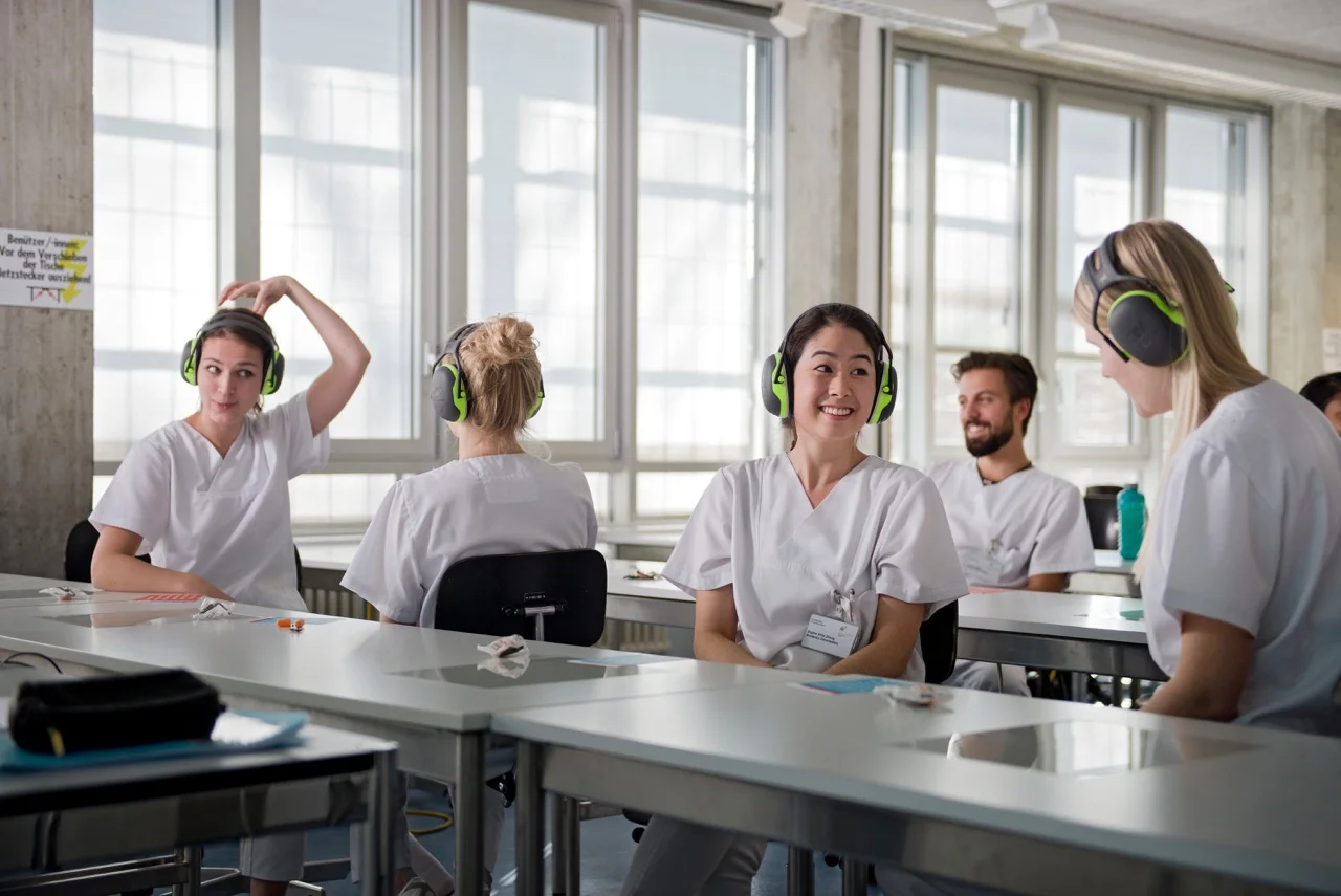 Studierende der zmk sitzen mit Gehörschützen im Vorlesungssaal und üben die Kommunikation bei Höreinschränkungen.