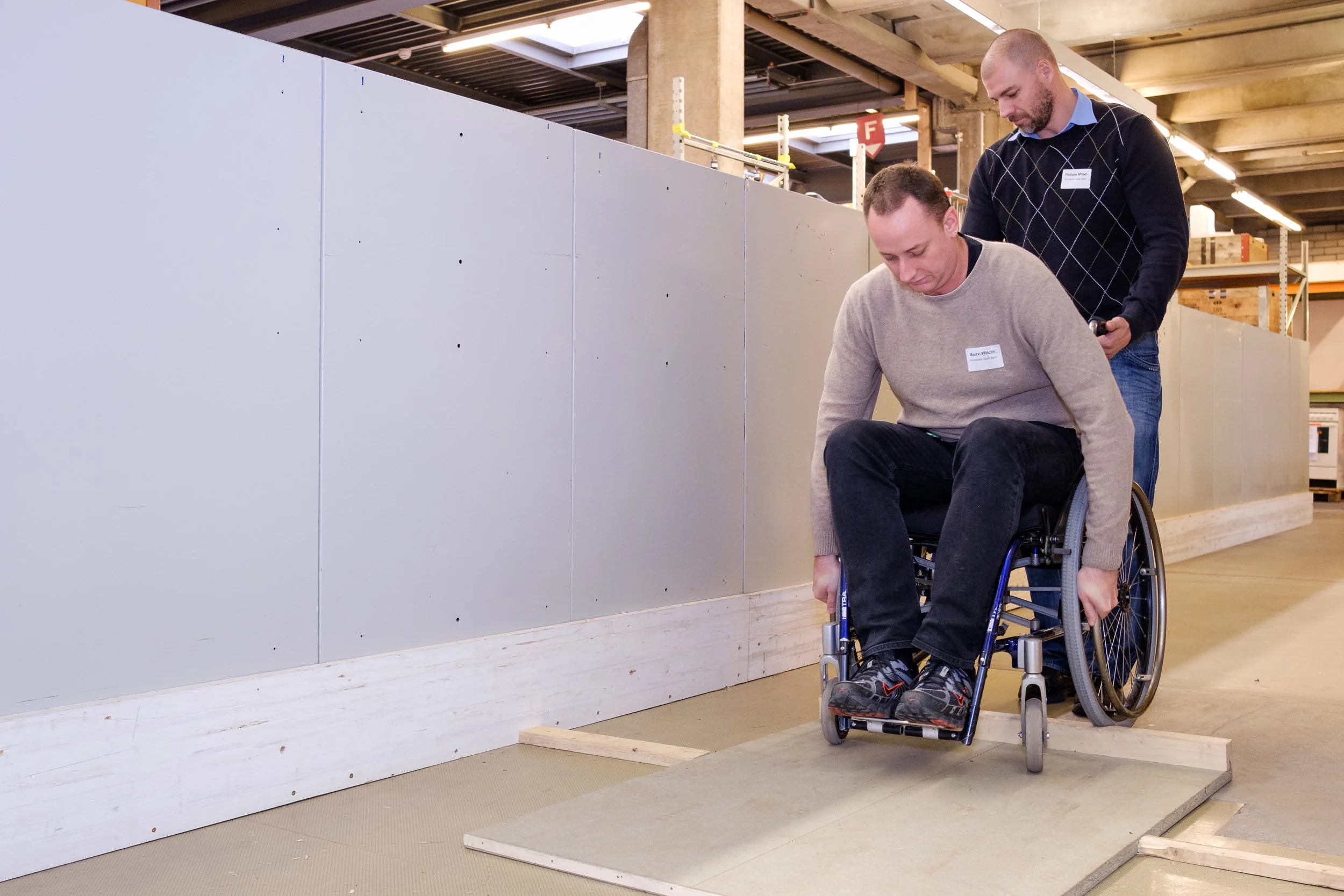 Ein Mann im Rollstuhl versucht vornübergebeugt über eine höhere Schwelle zu fahren. Dabei wird er von einem anderen Workshopteilnehmer gesichert.