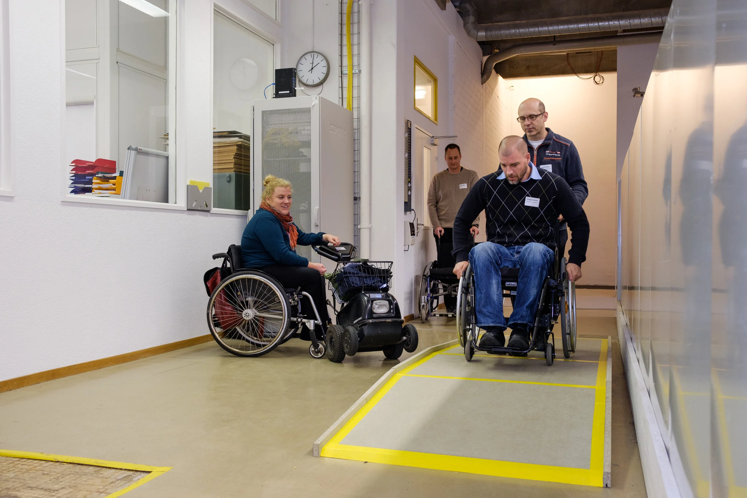 Ein Worshop-Teilnehmer im Rollstuhl fährt eine aufgebaute Rampe hoch und wird von seinem Kollegen hinten gesichert. Die Expertin im Rollstuhl schaut zu. Hinten wartet die nächste Gruppe auf die Übung.