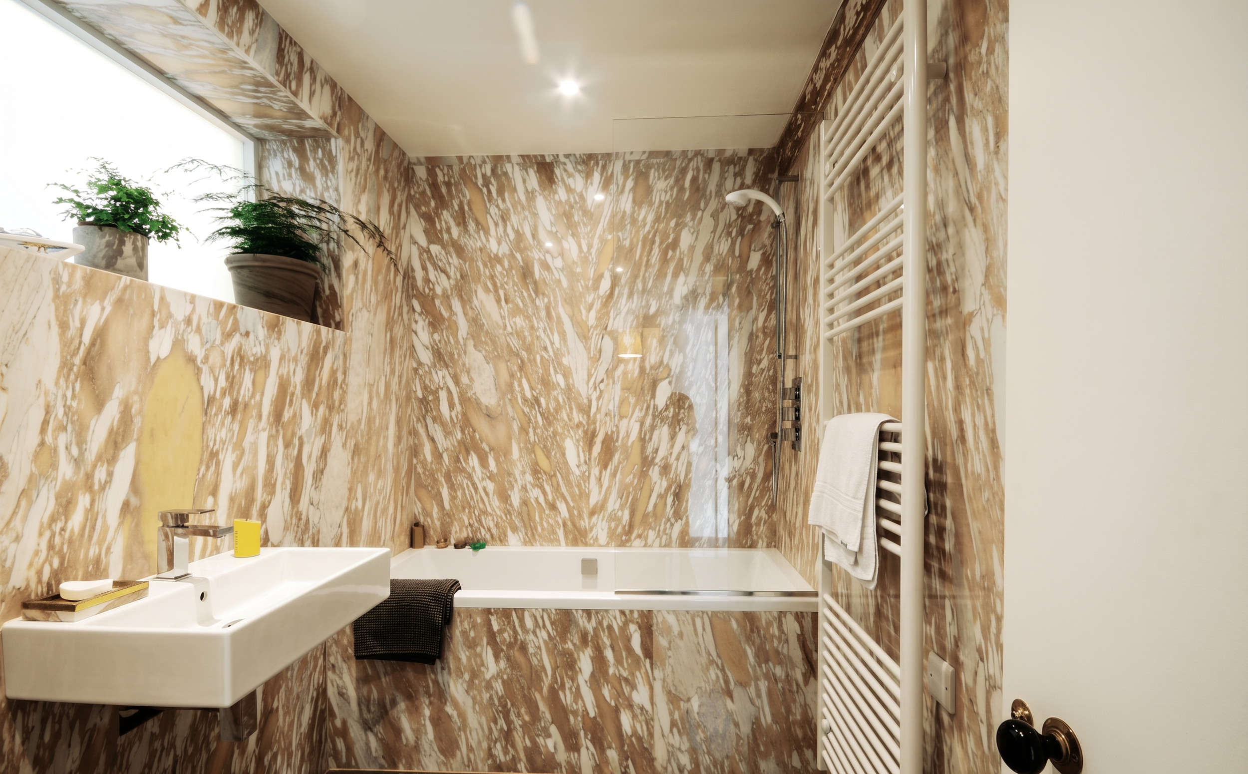 Bathroom with marble walls and ceiling, white sink, window with potted plants, towel radiator, and shower area.