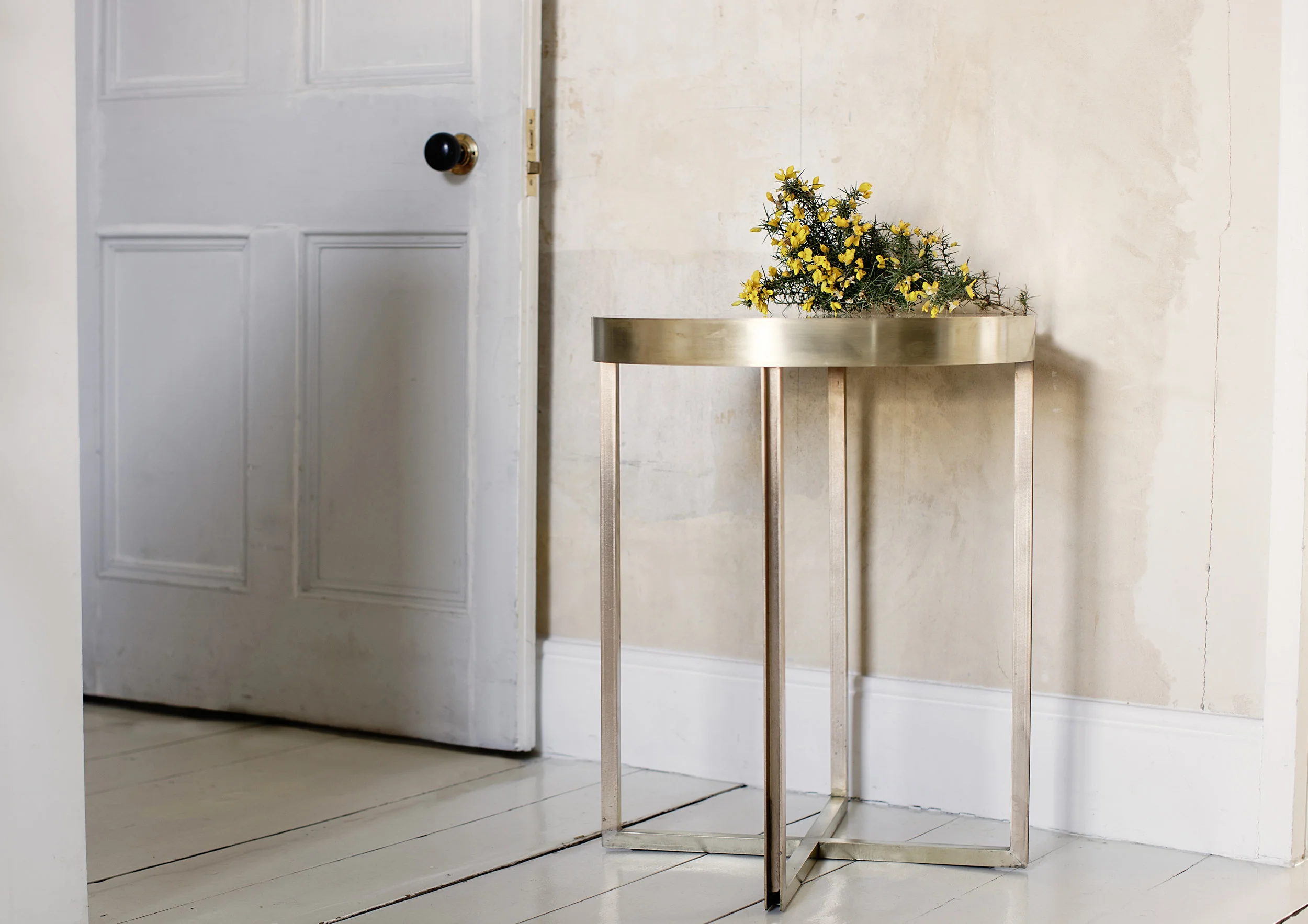 A metal table with a gold finish and four thin legs is positioned in front of a white door with black doorknob. There is a small bouquet of yellow flowers with green stems placed on the table. The background features a plain beige wall and a white ti