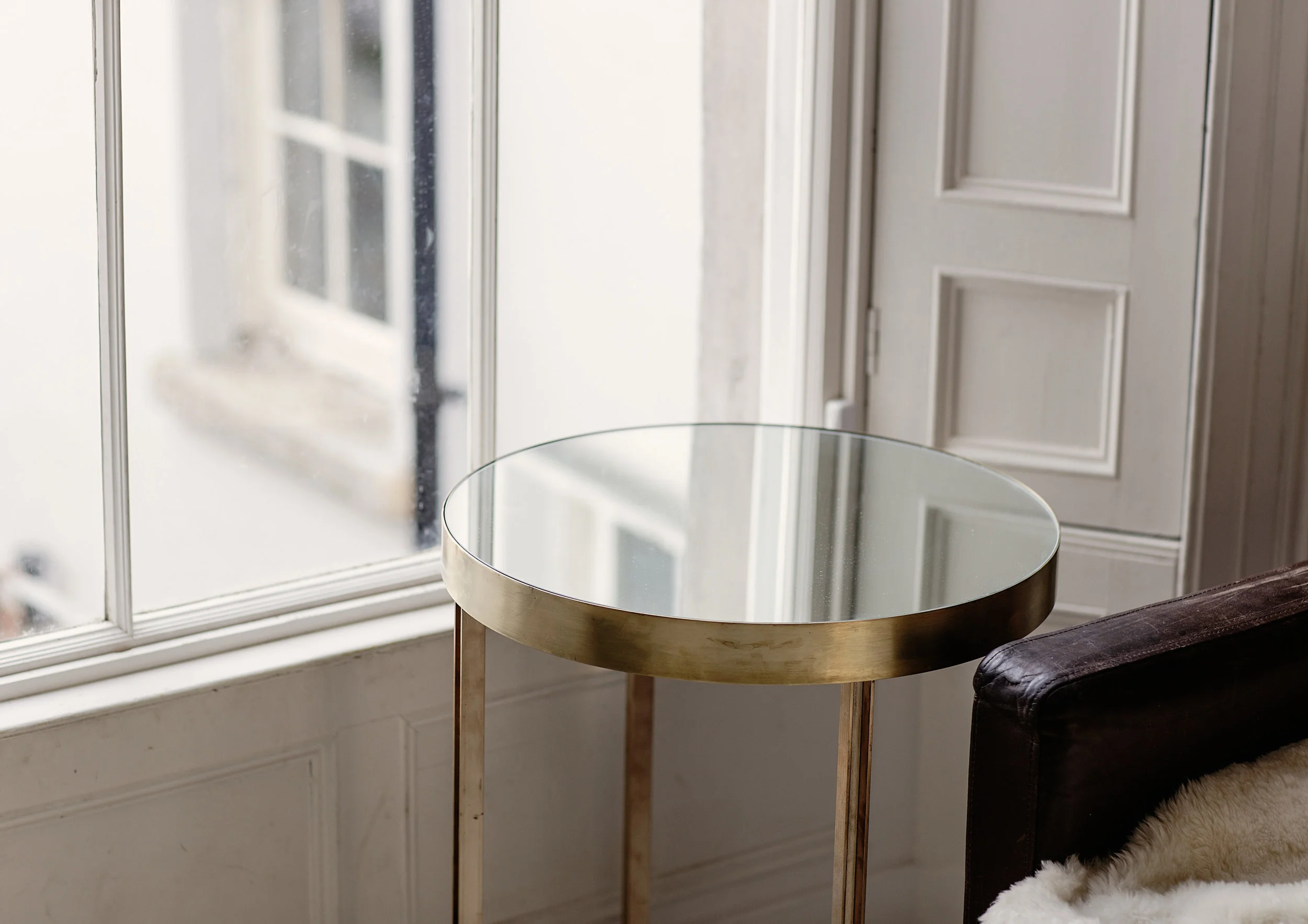 A round glass-topped side table with wooden legs placed next to a window with white trim. Part of a dark armchair and a white and black furry rug are also visible in the corner.